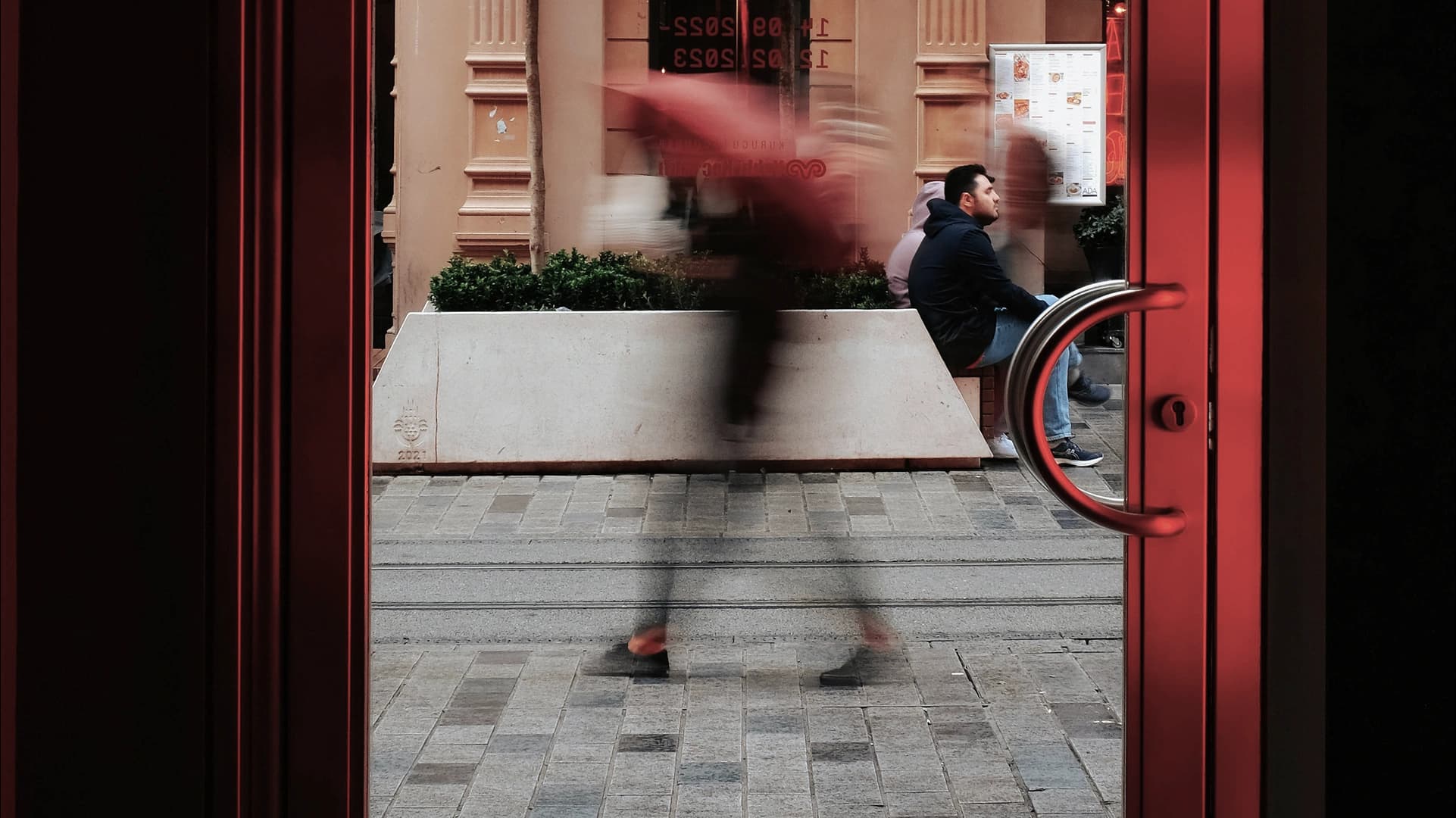 Blurred pedestrian walking past a seated man, viewed through red doorway.