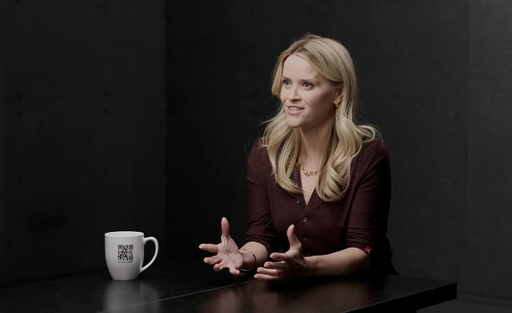 Reese Witherspoon speaking with coffee cup on table